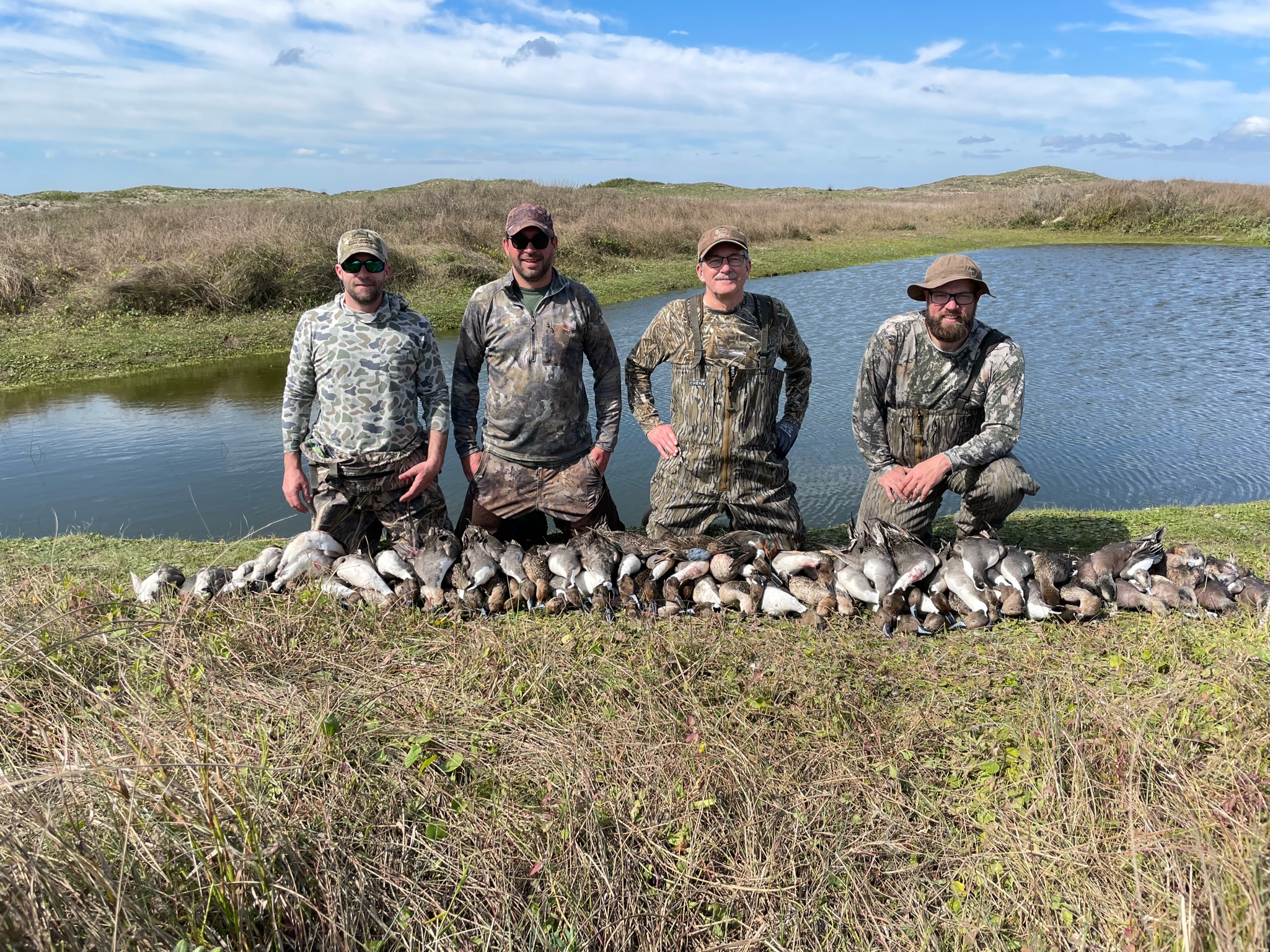 Hunters in camouflage gear posing with a successful duck hunting catch on the Laguna Madre, surrounded by wetlands and a pond, showcasing the excitement of wingshooting adventures in Mexico.