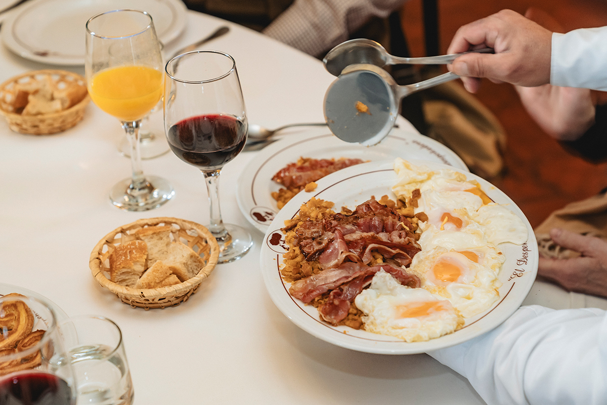 Plate of crispy bacon and fried eggs being served at a dining table, accompanied by glasses of red wine and orange juice, with a basket of bread in the background, highlighting a traditional Spanish meal experience.