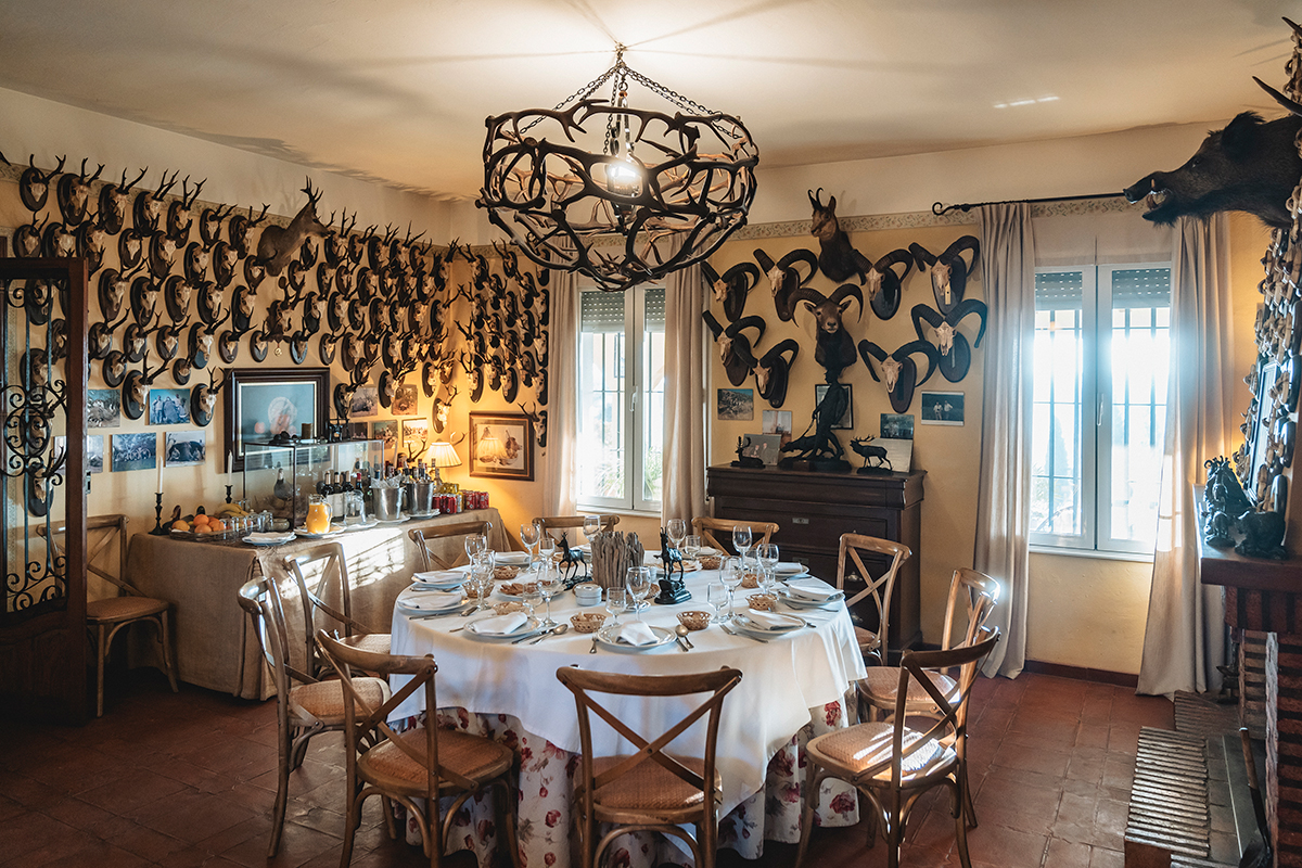 Dining room at El Despeñadero estate, featuring a round table set for a meal, surrounded by hunting trophies on the walls, and a rustic chandelier, reflecting Spain's wingshooting tradition.