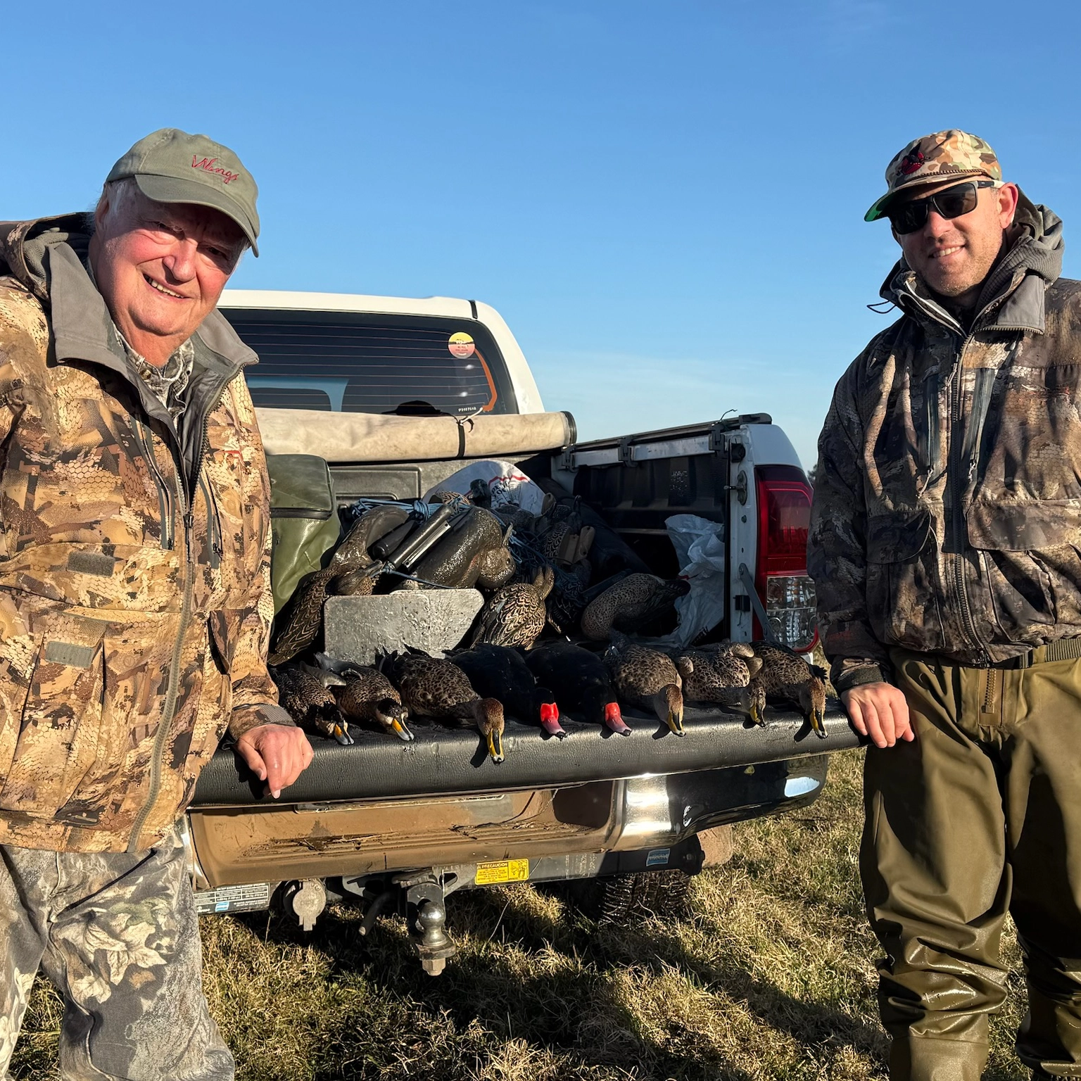 Men in camouflage attire standing by a pickup truck, showcasing their successful bird hunting trip with several ducks displayed on the tailgate, emphasizing the sporting lifestyle and hunting experience.