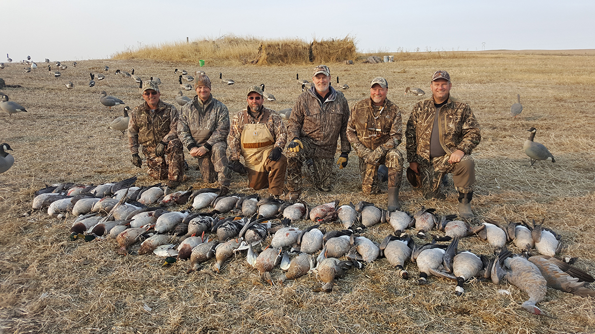 Group of hunters in camouflage gear posing with a successful waterfowl harvest, featuring various ducks and geese laid out in front of them on a prairie landscape, with additional birds in the background.