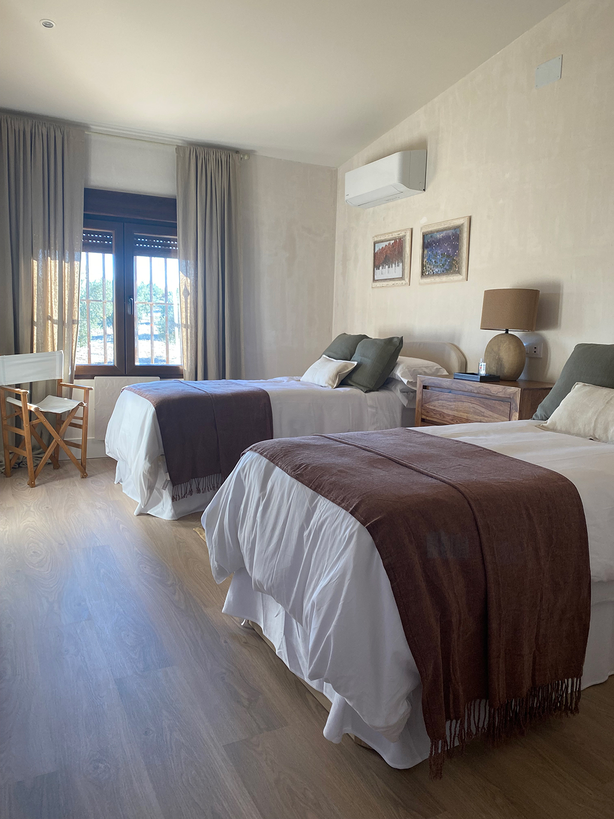 Spacious lodge room at El Despeñadero featuring two beds with white linens and brown throws, elegant decor, large window with natural light, and wooden furniture, ideal for wingshooting guests in Spain.