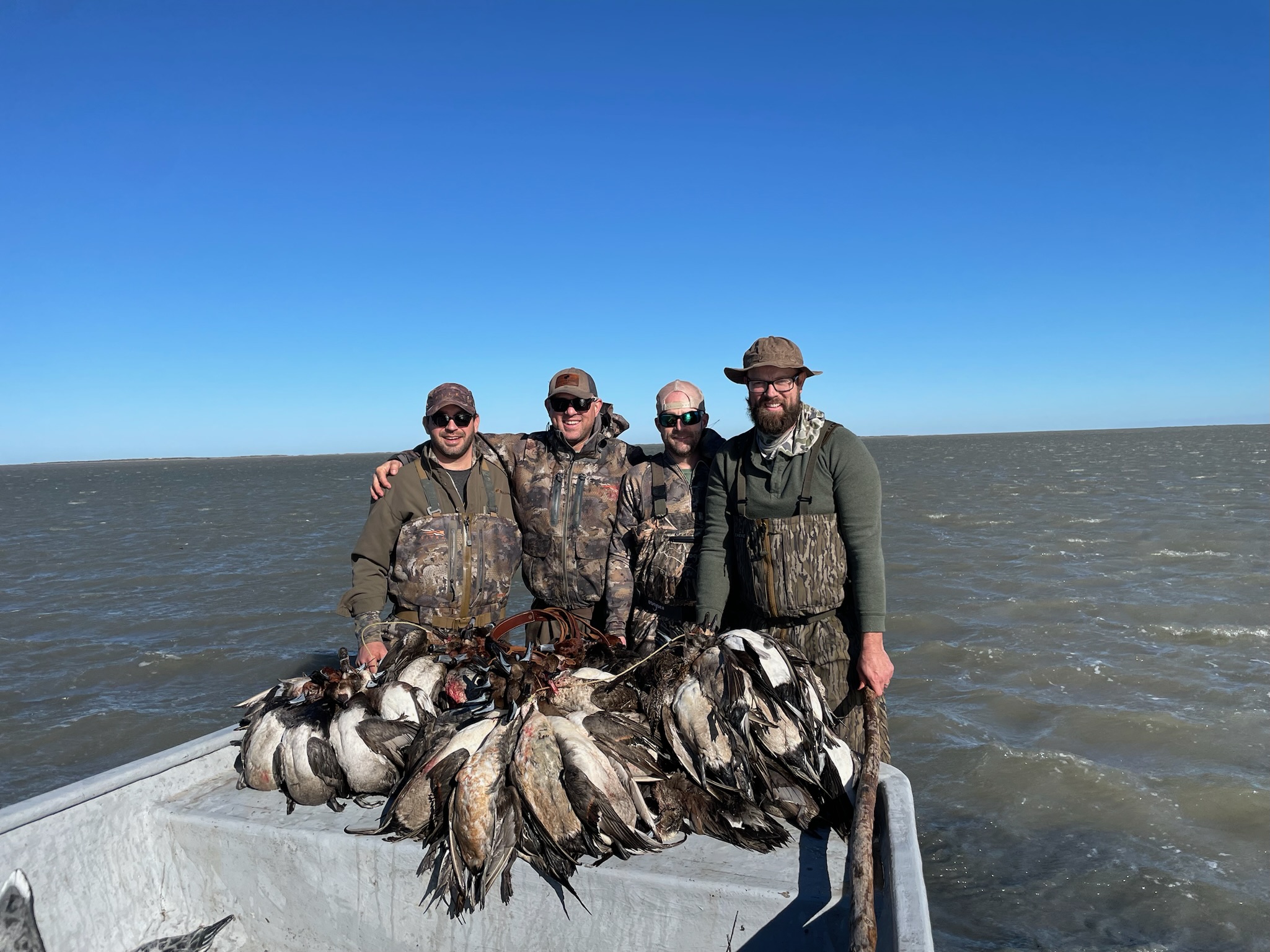 Group of hunters in camouflage gear posing on a boat with a successful duck hunting catch, featuring a variety of ducks, against the backdrop of the Laguna Madre.