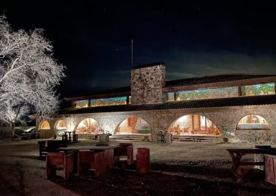 El Despeñadero hunting lodge at night, showcasing stone architecture, illuminated windows, and outdoor seating, set against a dark sky, representing a premier wingshooting destination in Spain.