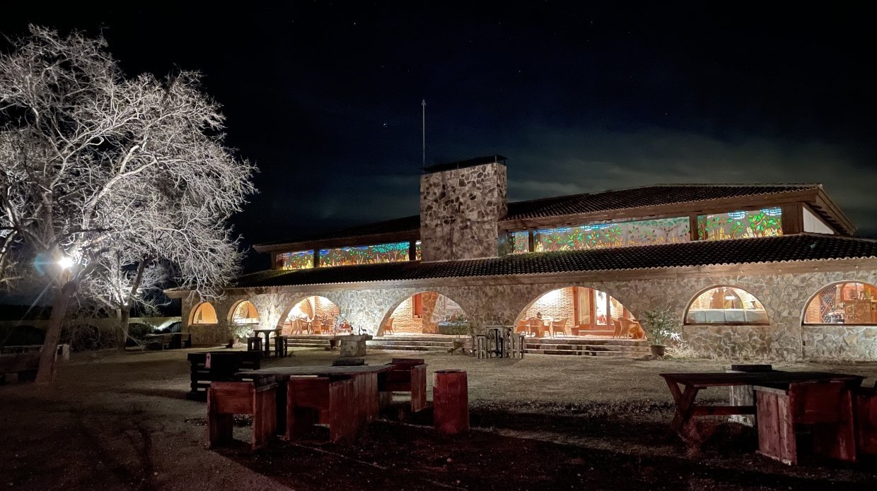 El Despeñadero hunting lodge at night, showcasing stone architecture, illuminated windows, and outdoor seating, set against a dark sky, representing a premier wingshooting destination in Spain.