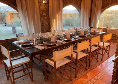 Elegant dining setup at El Despeñadero Lodge, featuring a long wooden table set for a meal, surrounded by rustic stone walls and large windows overlooking the estate's scenic landscape.