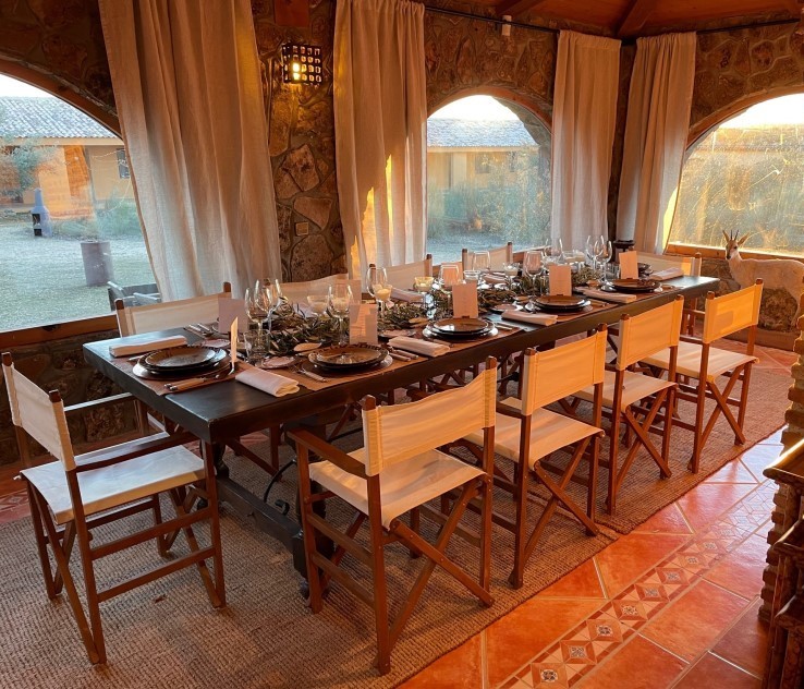 Elegant dining setup at El Despeñadero Lodge, featuring a long wooden table set for a meal, surrounded by rustic stone walls and large windows overlooking the estate's scenic landscape.