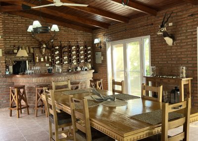 Interior view of Estancia La Emilia dining area featuring rustic wooden furniture, a bar with wine bottles, and traditional Argentine decor, highlighting the luxurious accommodations for hunting guests.