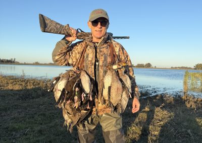 Hunter in camouflage holding a shotgun and a bag of ducks, standing by a lagoon in La Paz, Argentina, showcasing a successful duck hunting experience.