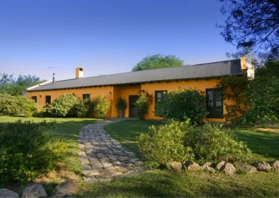 Lodge exterior of Las Vertientes in Córdoba, Argentina, featuring a yellow building surrounded by lush greenery and a stone pathway, reflecting the relaxed atmosphere for wingshooting adventures.