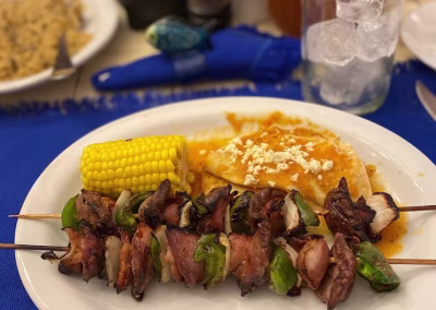 Plate of grilled meat skewers with bell peppers, served with corn on the cob and cheese-topped enchiladas, reflecting the authentic dining experience at La Marina Del Río lodge in Mexico.