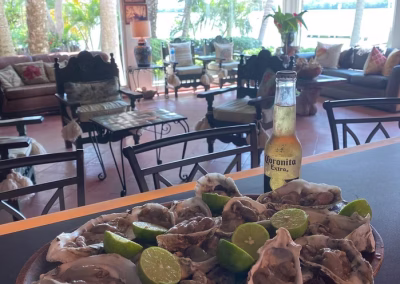 Plate of oysters with lime and a Corona beer in foreground, cozy lodge interior with comfortable seating and scenic view of the outdoors in background, highlighting La Marina del Río's inviting atmosphere for guests after a day of hunting.