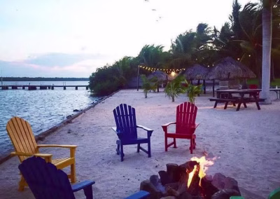 Colorful Adirondack chairs by a beachside fire pit at La Marina Del Río, with palm trees and a pier in the background, highlighting a relaxing atmosphere for guests after hunting adventures.
