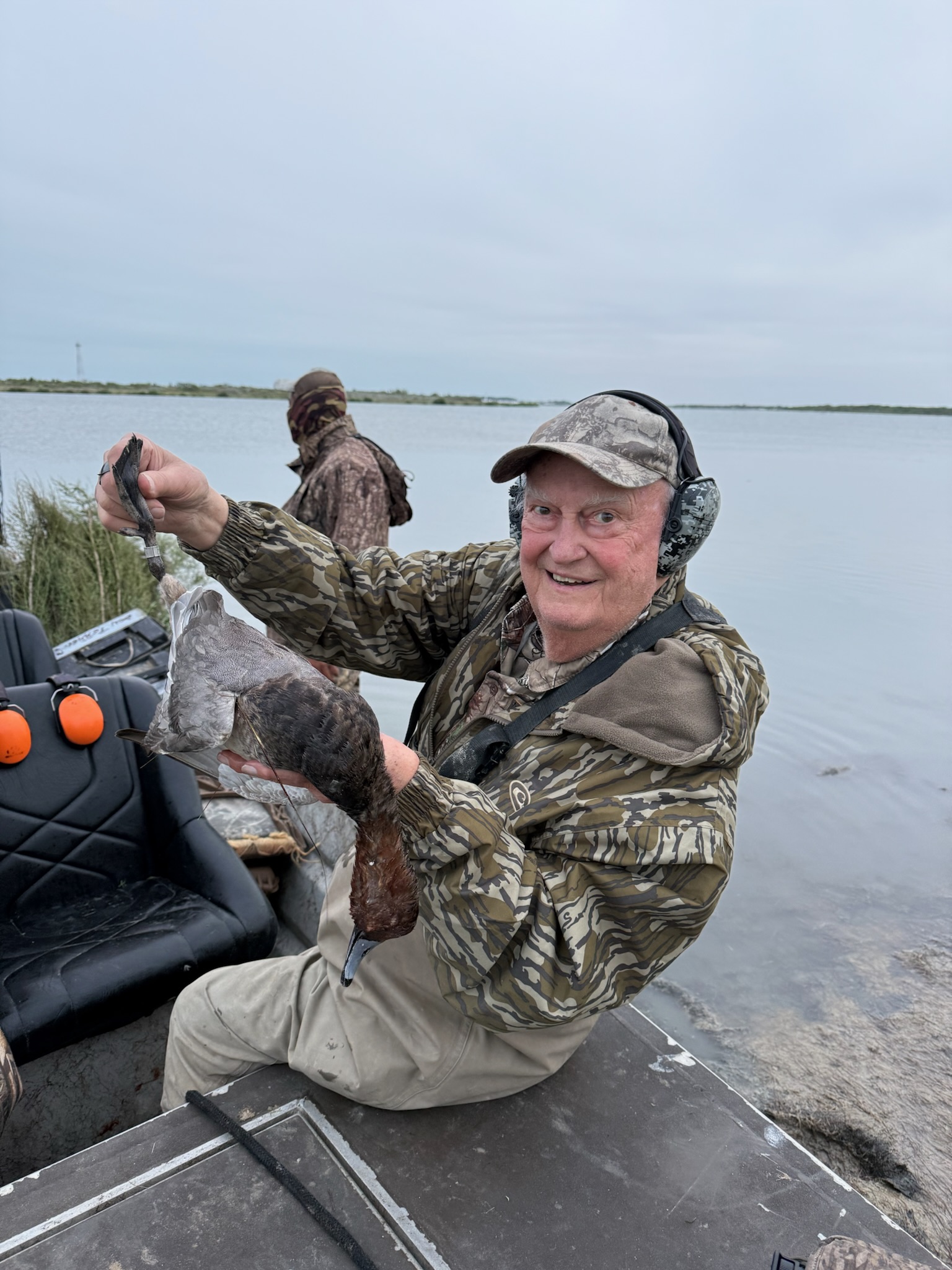 Smiling man in camouflage clothing holding a duck in a hunting boat on the Laguna Madre, showcasing a successful wingshooting experience in Mexico.