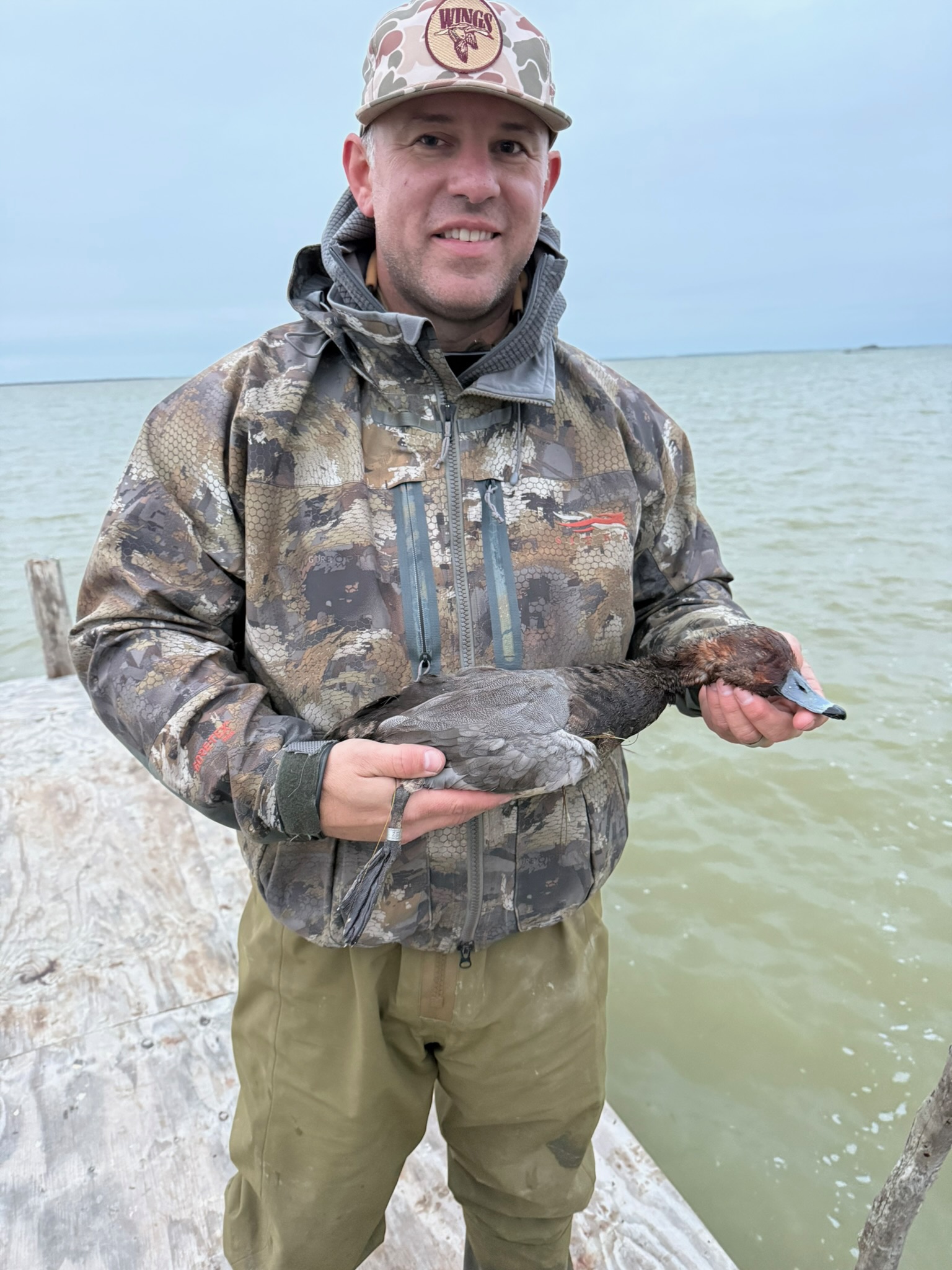Man in camouflage holding a duck, standing on a dock by the Laguna Madre, showcasing wingshooting adventure in Mexico.