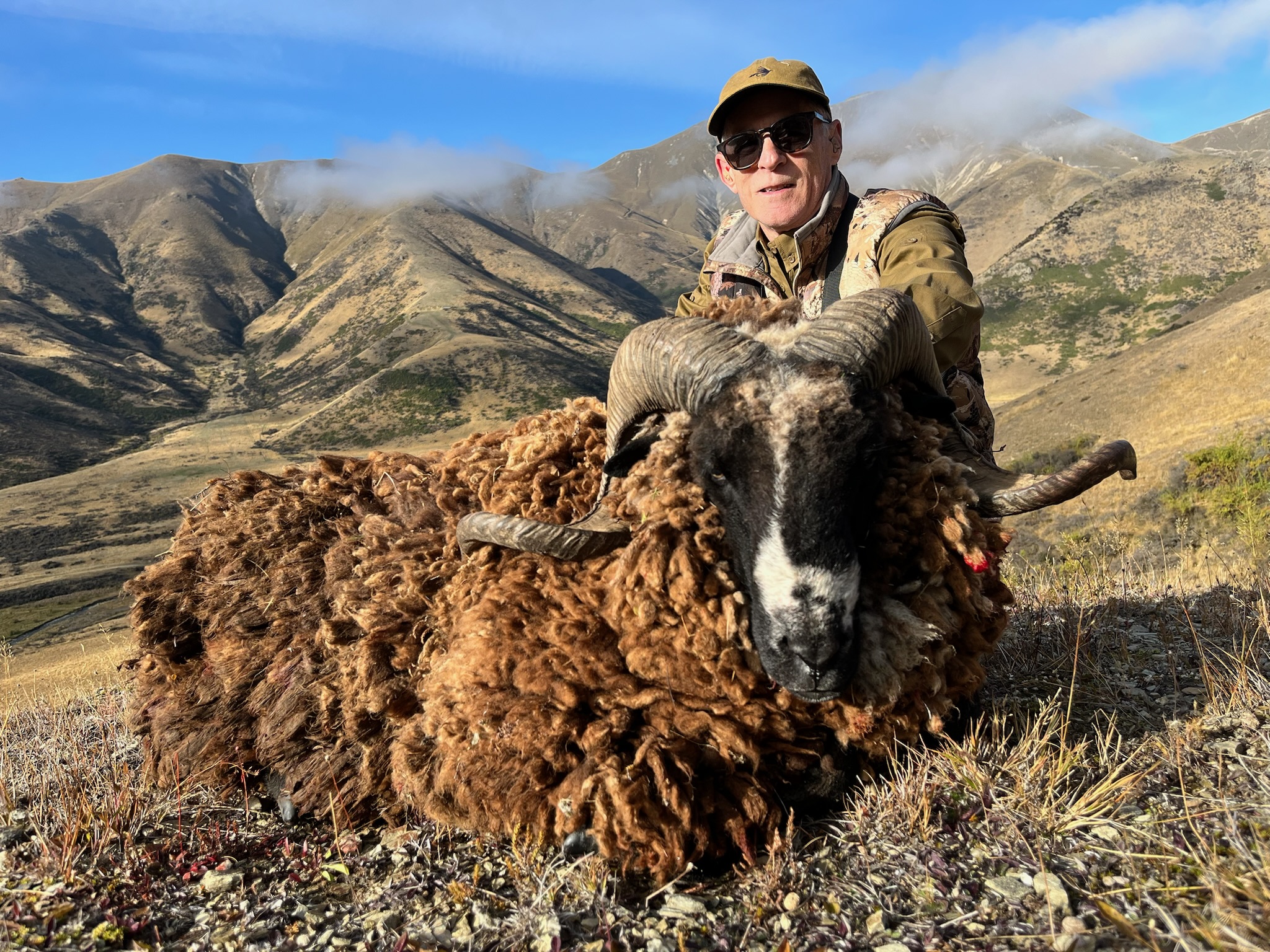 Hunter posing with a trophy ram in a mountainous New Zealand landscape, showcasing the region's exotic big game hunting opportunities.