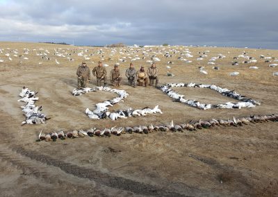 Hunters posing in front of a display of harvested snow geese and ducks arranged in the shape of "120," set against a wide-open prairie landscape, highlighting a successful waterfowl hunting experience in Saskatchewan.