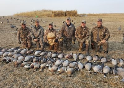 Hunters posing in camouflage gear with a successful waterfowl harvest, featuring various species of geese, on a Saskatchewan prairie landscape.