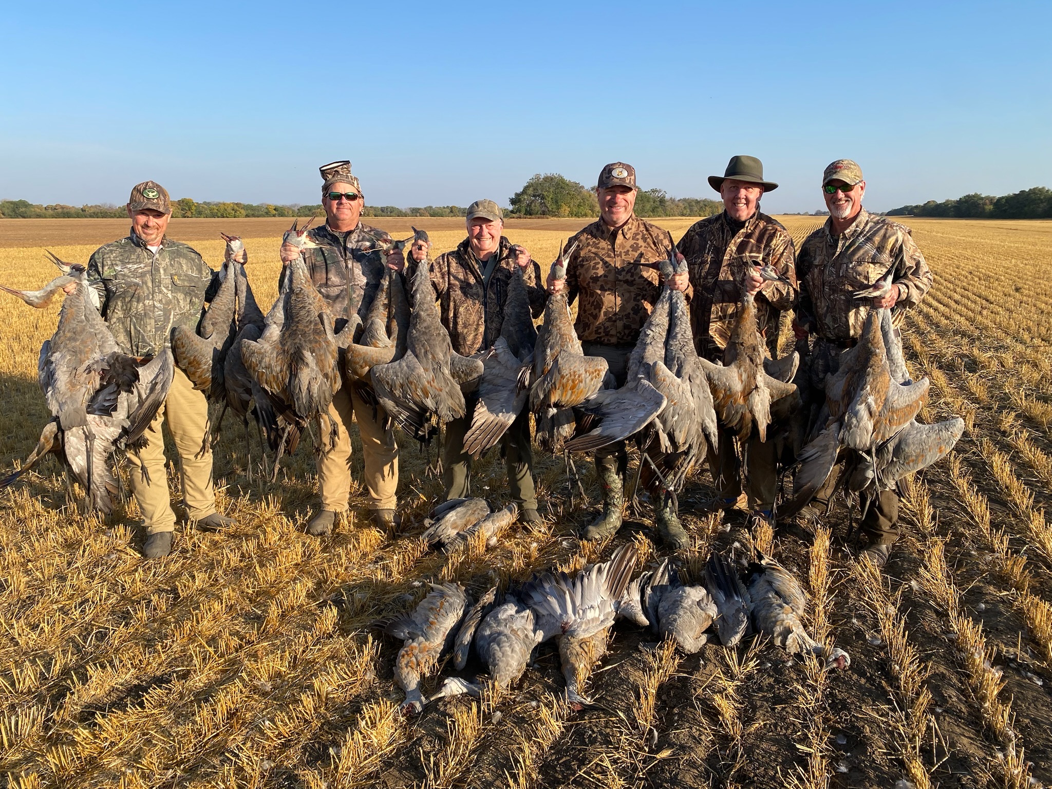 Wings-Saskatchewan-featured Group of hunters in camouflage attire holding harvested sandhill cranes in a Saskatchewan grain field, showcasing successful waterfowl hunting experience.