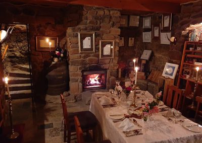 Cozy dining area with a stone fireplace, elegantly set table featuring floral centerpieces and fine tableware, surrounded by rustic decor and framed certificates.