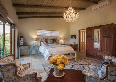 Elegant bedroom with wooden beams, chandelier, and stylish furnishings, featuring a king-sized bed, decorative pillows, and a cozy seating area with patterned chairs, ideal for relaxing after a South Africa wingshooting safari.