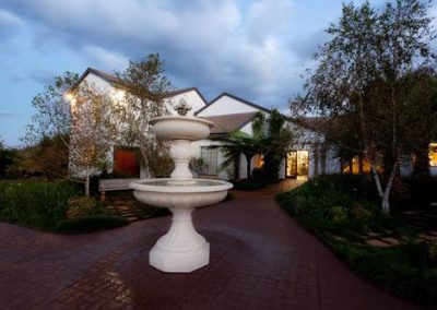 Elegant entrance of a South African hunting lodge featuring a central white fountain, surrounded by lush greenery and modern architecture, emphasizing the serene setting for wingshooting adventures.