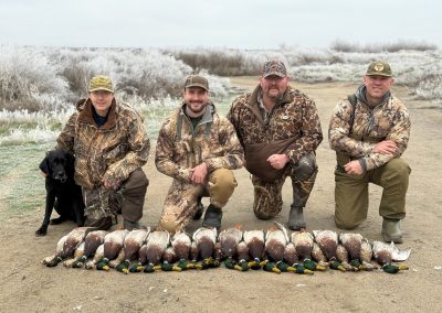 Hunters in camouflage gear posing with a successful mallard duck hunt, showcasing a row of harvested ducks on a sandy area, emphasizing the guided hunting experience offered by Pacific Wings in the Columbia Basin.