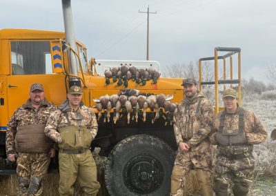 Four hunters in camouflage gear standing in front of a large truck displaying a successful mallard hunt, showcasing their harvest of ducks, with a rural backdrop typical of the Columbia Basin hunting environment.