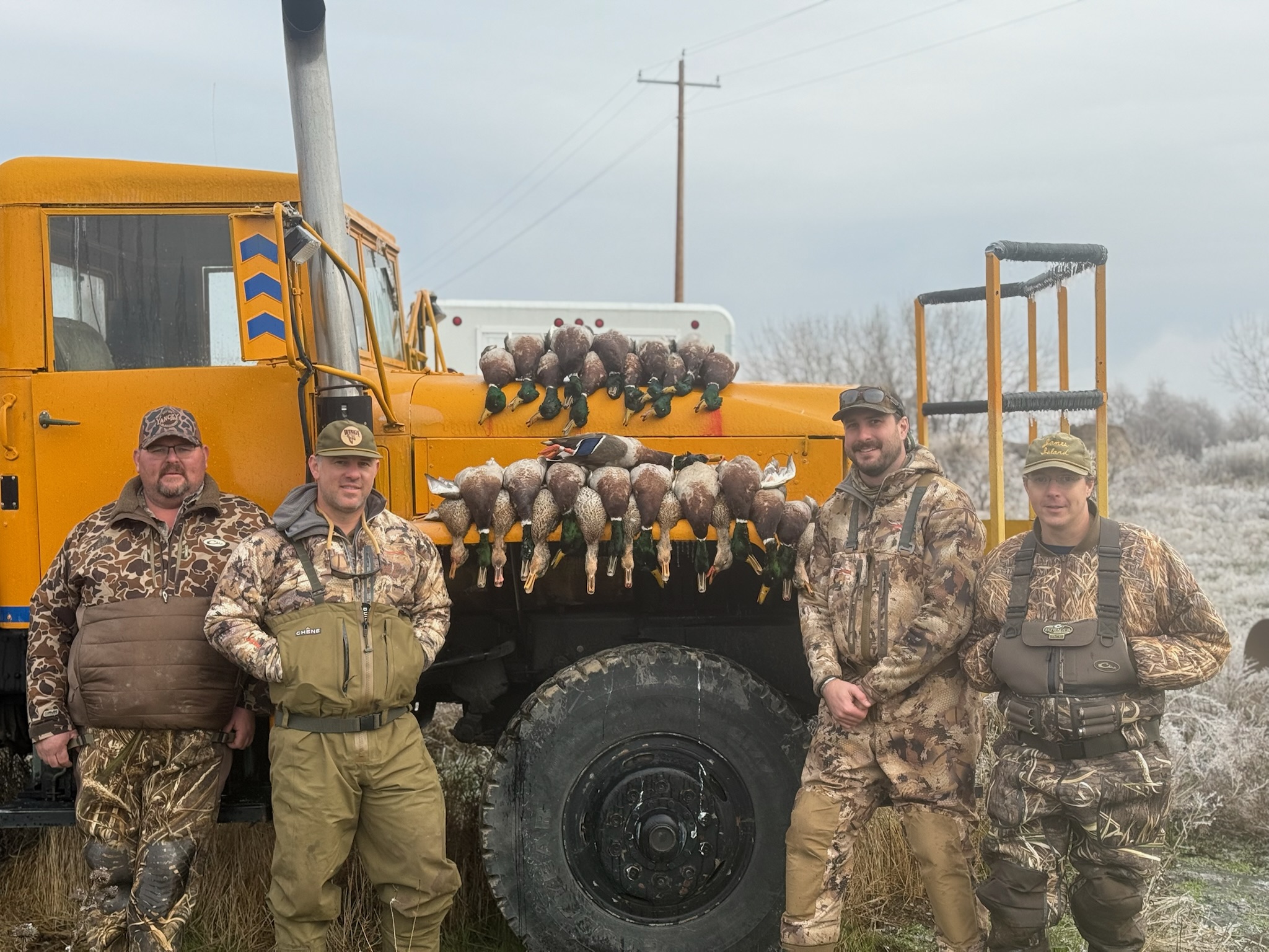 Hunters in camouflage gear posing next to an orange vehicle, showcasing a successful mallard duck hunt with several ducks displayed on the vehicle's front.