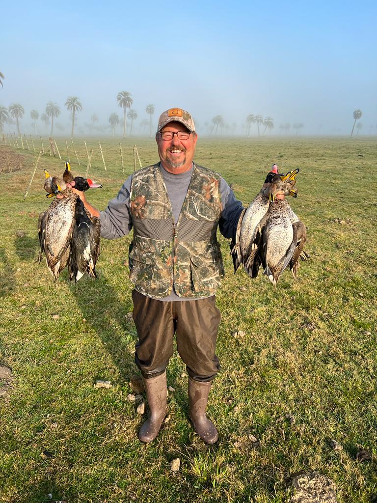 Wings-Uruguay-11 Hunter proudly displaying harvested ducks in Uruguay's wetlands, showcasing a successful duck hunting experience amidst palm trees and mist.