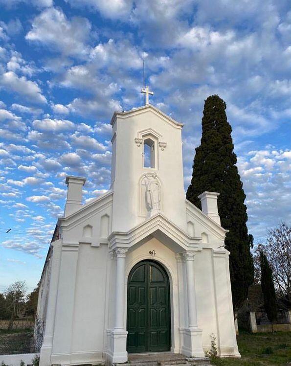 Wings-Uruguay-13 White chapel with green door and cross, surrounded by trees and blue sky with clouds, representing Uruguay's serene landscapes near duck hunting and perdiz shooting destinations.