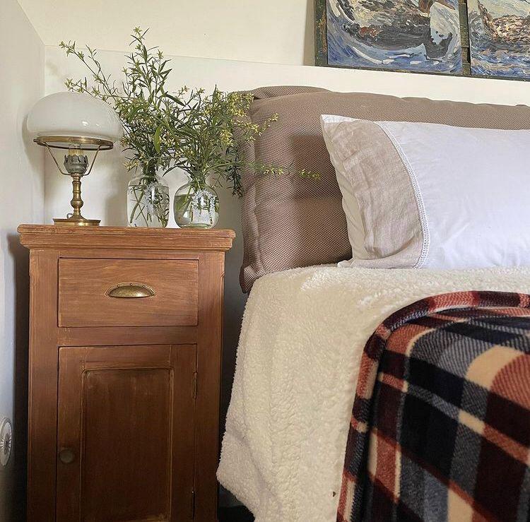 Cozy bedroom corner featuring a wooden nightstand with a brass lamp, two glass vases with greenery, a plush white blanket, and a patterned throw, creating a serene and inviting atmosphere.