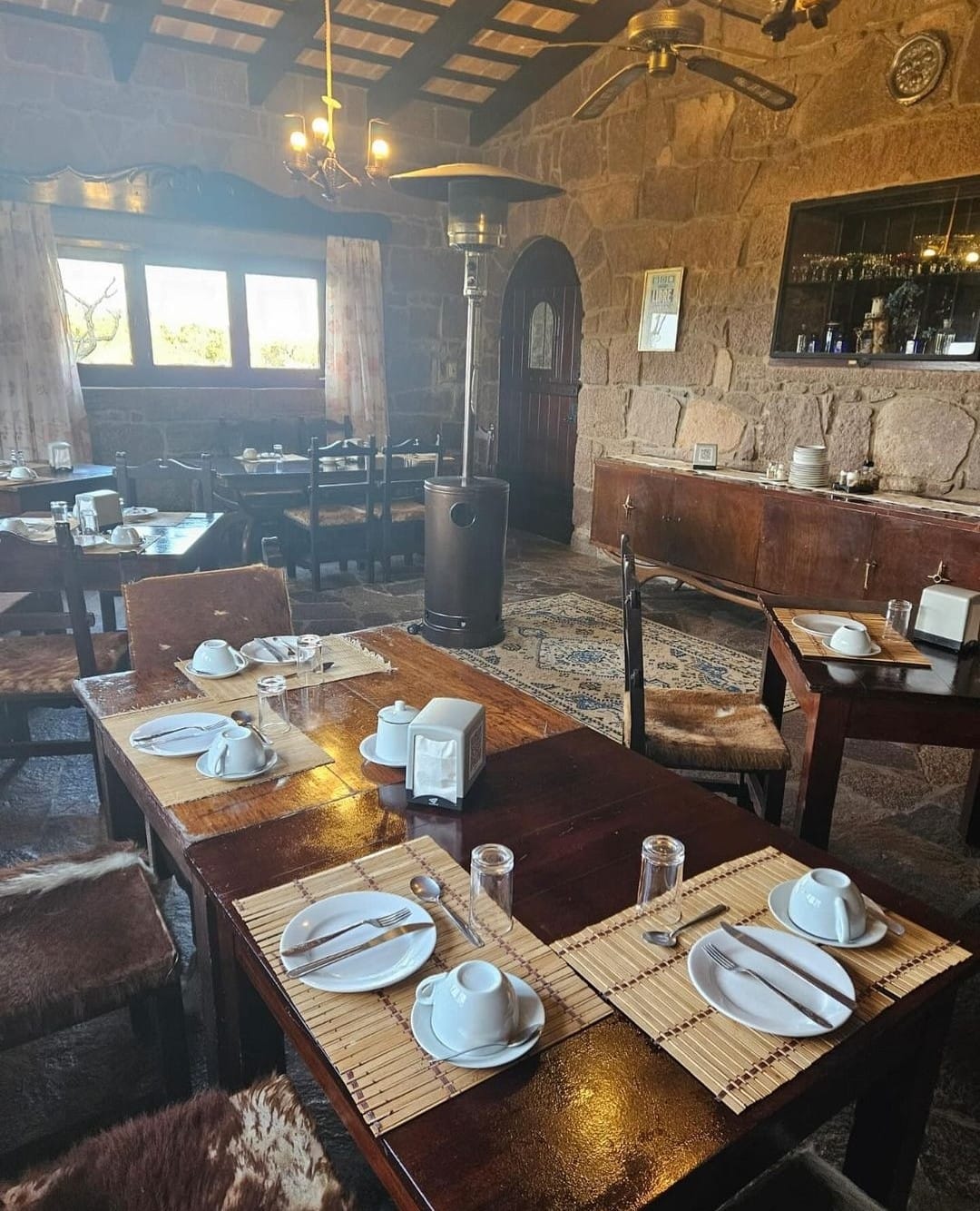 Rustic dining area in a lodge featuring wooden tables set with plates, cutlery, and glassware, with a cozy ambiance enhanced by a heater and stone walls, ideal for post-hunt meals in Uruguay.