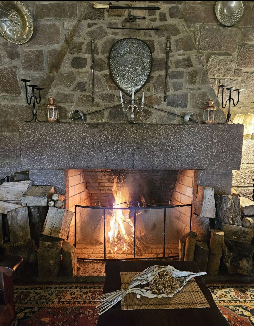 Cozy fireplace with a stone surround, decorative shields and weapons on the wall, and a rustic wooden table with a decorative bundle, enhancing the warm atmosphere of the lodge in Uruguay.